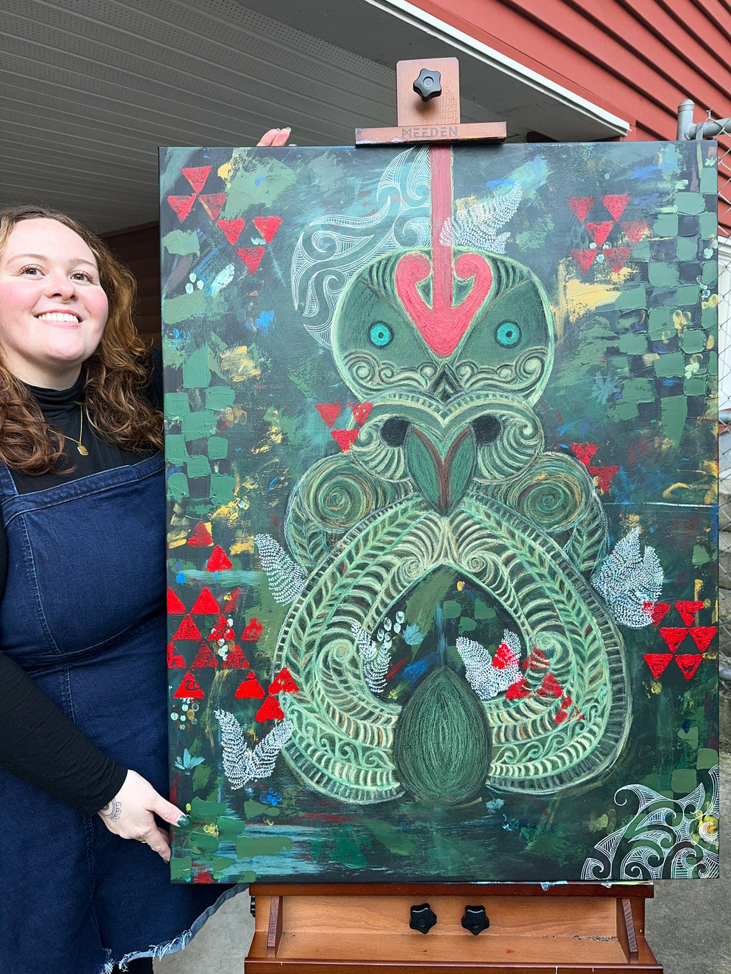 Person holding a colorful painting of a stylized green tiki with red triangles on an easel.